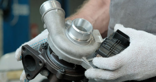 Gloved hands holding a silver and black turbocharger part in a car service area with blurred equipment behind.