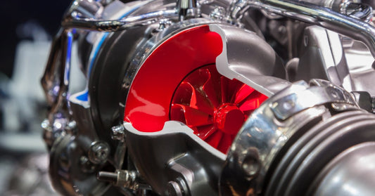 A cutaway turbocharger with a red turbine wheel inside gray housing, surrounded by shiny metal pipes and bolts.