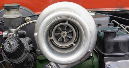 A large silver turbocharger mounted among hoses and engine parts beneath a red car body panel and surrounding black parts.