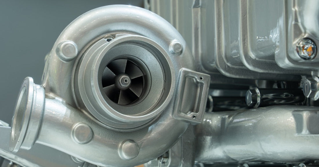 A silver turbocharger with dark turbine blades sits beside a metal engine block with bolts and ribbed panels.