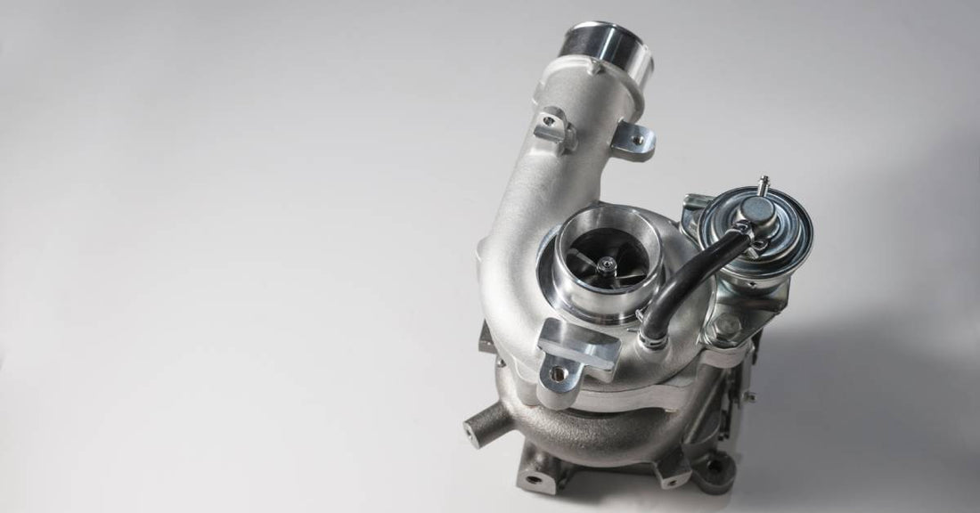 A shiny silver turbocharger sits upright on top of a plain white surface, casting a shadow to the right.