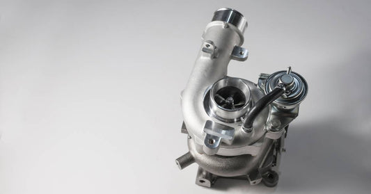 A shiny silver turbocharger sits upright on top of a plain white surface, casting a shadow to the right.