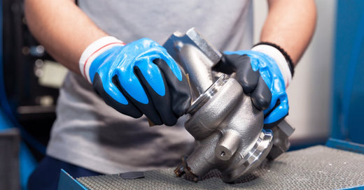 A person wearing blue gloves and a gray shirt handles a metallic turbocharger component on a workbench in a workshop setting.