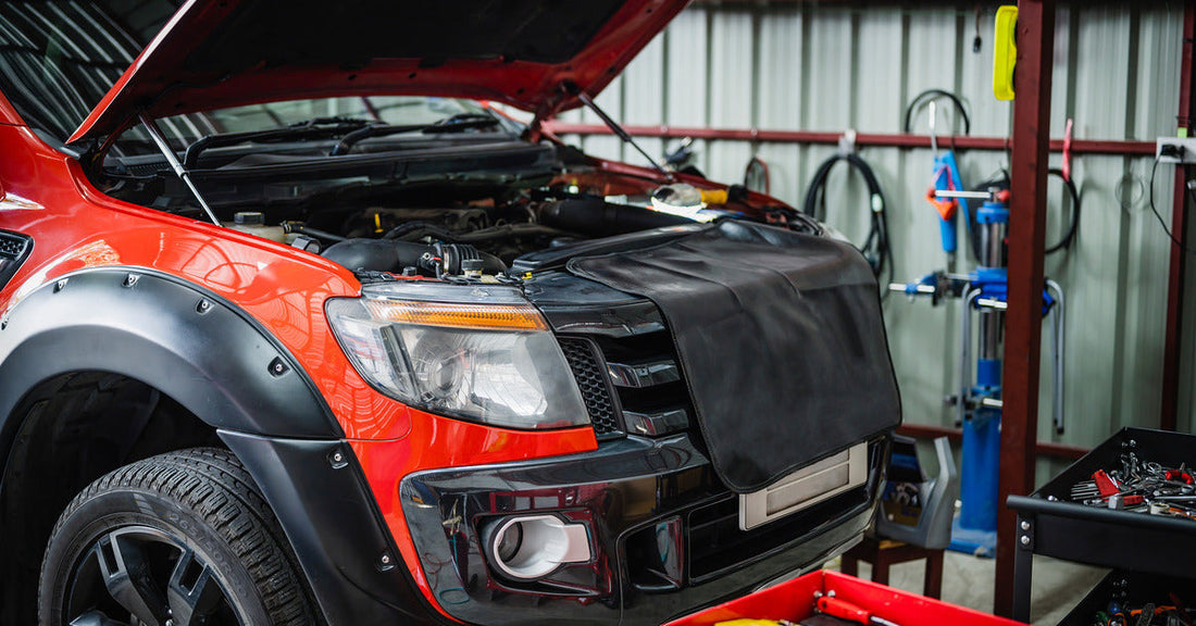 A red truck parked in a garage. The truck's hood is open, there are tools in a red cart nearby, and equipment hangs on the wall.