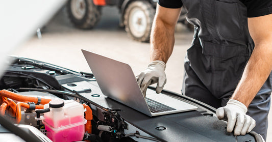 A mechanic wearing gloves and a gray jumpsuit works on a car while using a laptop placed on the engine bay.