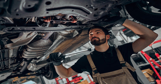 A male mechanic is examining the underbody of a car that is lifted up while holding a work light.
