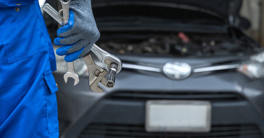 A close-up of a mechanic in blue overalls holding tools while a gray car is parked in the background.