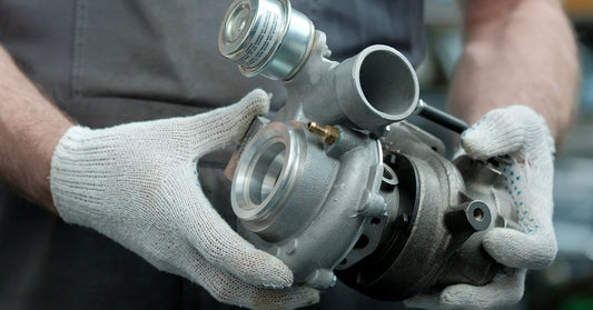 A mechanic wearing white gloves and a gray shirt is holding a brand-new, metallic turbocharger for a car.