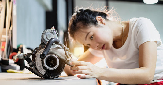 A woman wearing a white shirt and a necklace inspects a turbocharger sitting on top of a gray desk in a garage.
