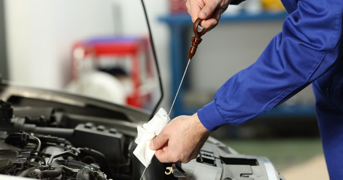 A mechanic wearing navy blue overalls uses a dipstick and a piece of cloth to check the oil level of a vehicle.
