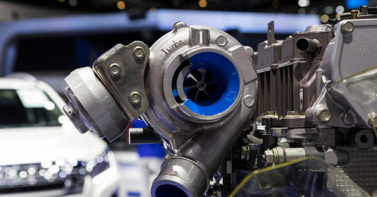 A turbocharger with a blue-lined turbine is attached to a diesel engine, with a white vehicle in the background.
