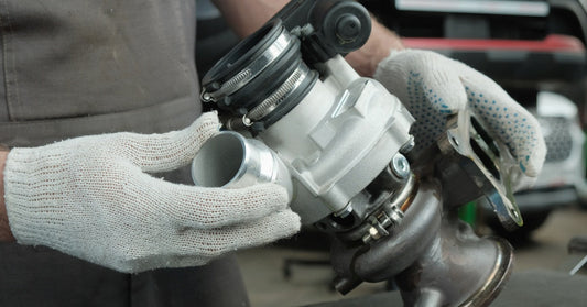 A person wearing white gloves holds and inspects a sleek, new turbocharger part placed on a black table.
