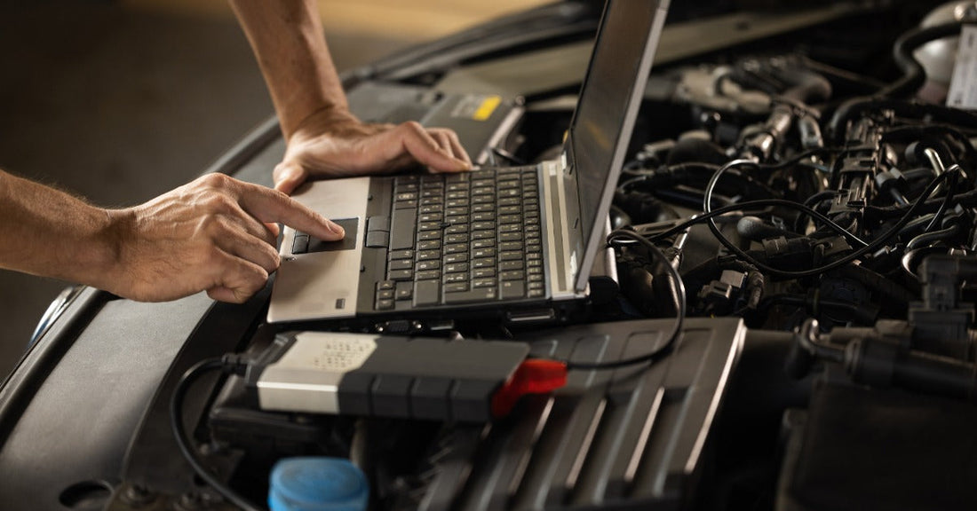 Mechanic uses a laptop beside an open car hood while inspecting vehicle systems during a diagnostic test.