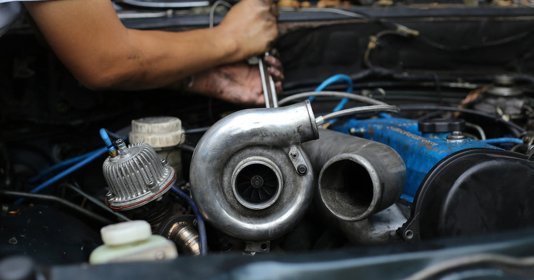 A close-up of a person using a hand tool to work on a car's engine. In front of the person is a turbocharger.