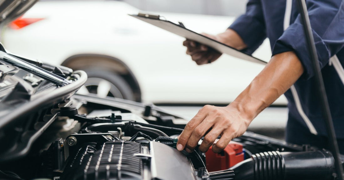 A person with a clipboard in their hand is inspecting a vehicle’s engine. They are touching the engine with their other hand.
