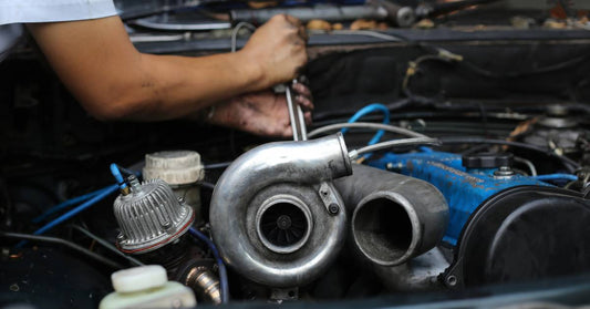 A man wearing short sleeves bent over the open hood of a car working on a turbocharged engine with tools.
