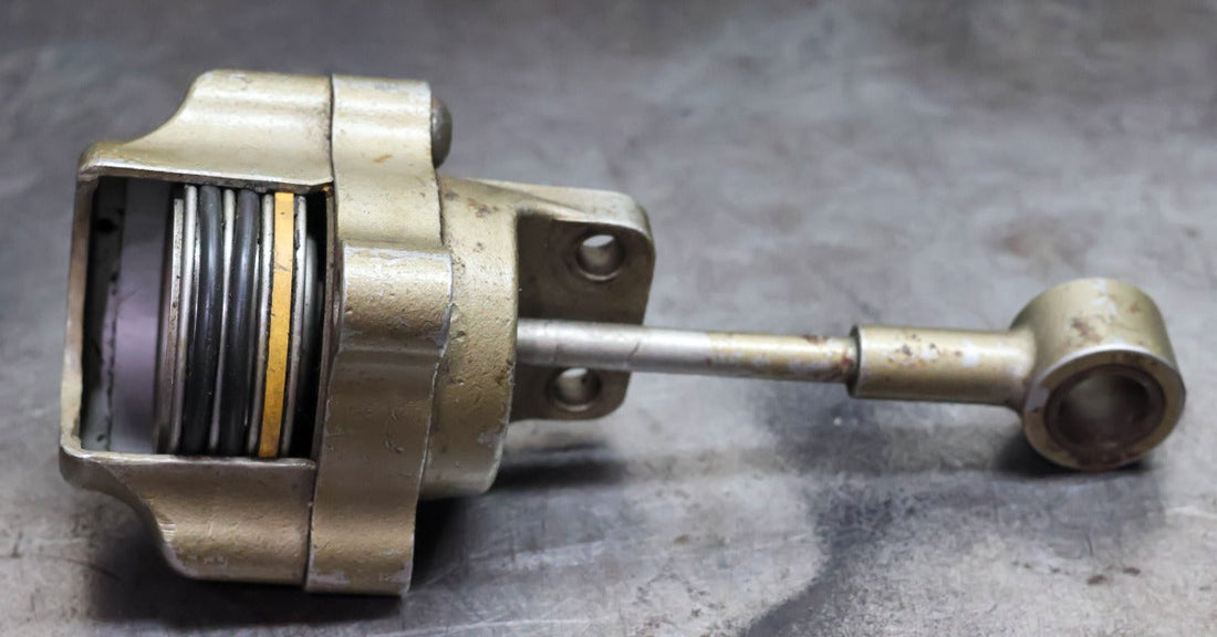 A cutaway metal actuator with a visible spring, round housing, and long rod resting on a scratched metal surface.