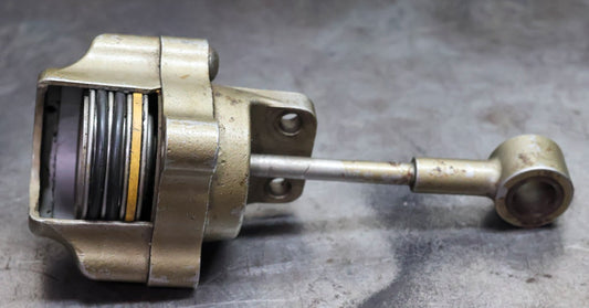 A cutaway metal actuator with a visible spring, round housing, and long rod resting on a scratched metal surface.