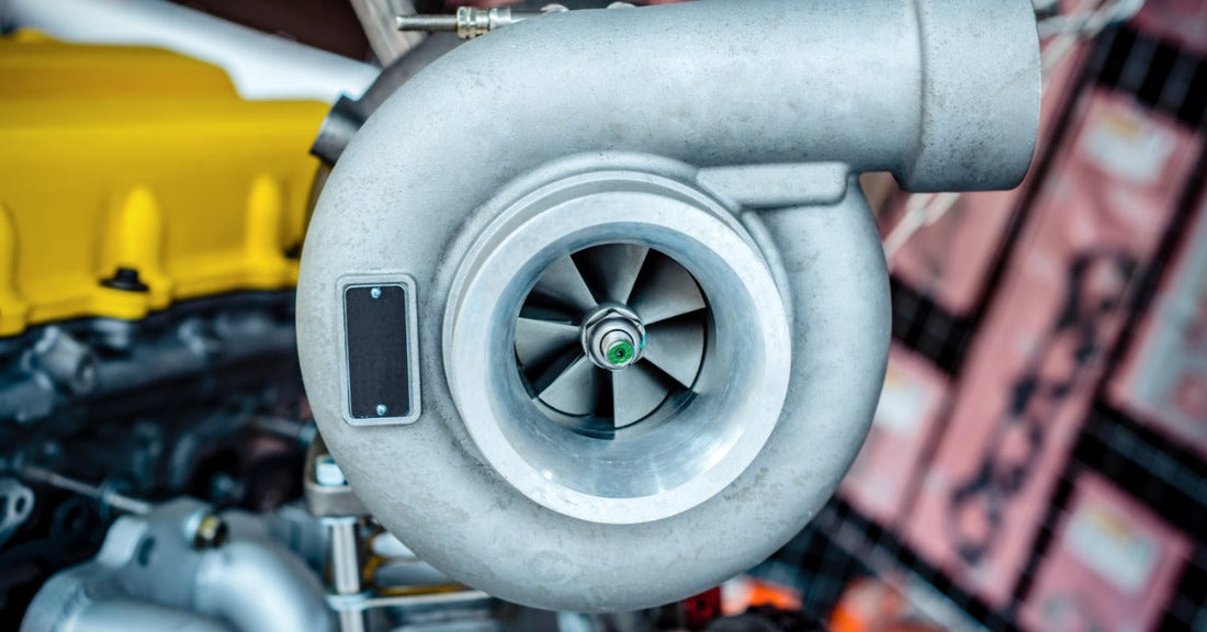 a close-up turbocharger with polished metal housing, curved pipes, bolts, and fittings in an automotive workshop.
