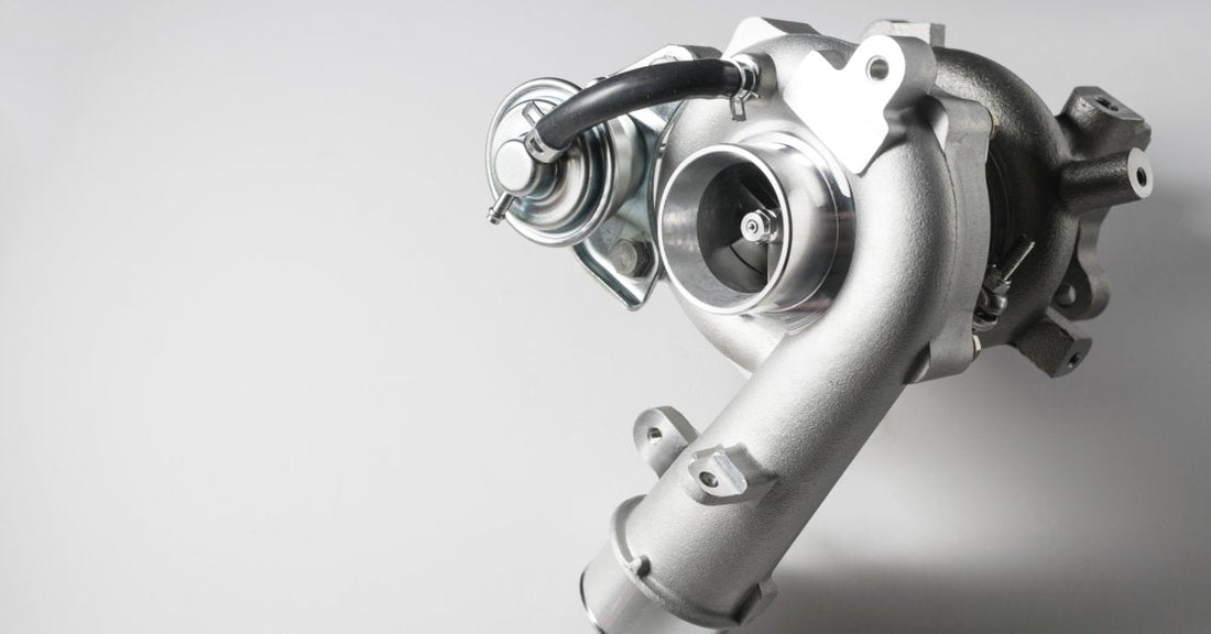 A silver and black turbocharger with a curved metal housing and attached hose on a light gray background.