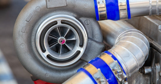 A close-up view of a metal turbocharger mounted on a car engine with hoses and automotive components visible.