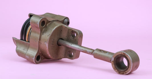 a weathered metal turbocharger actuator with an exposed spring and long rod resting against a pink background.