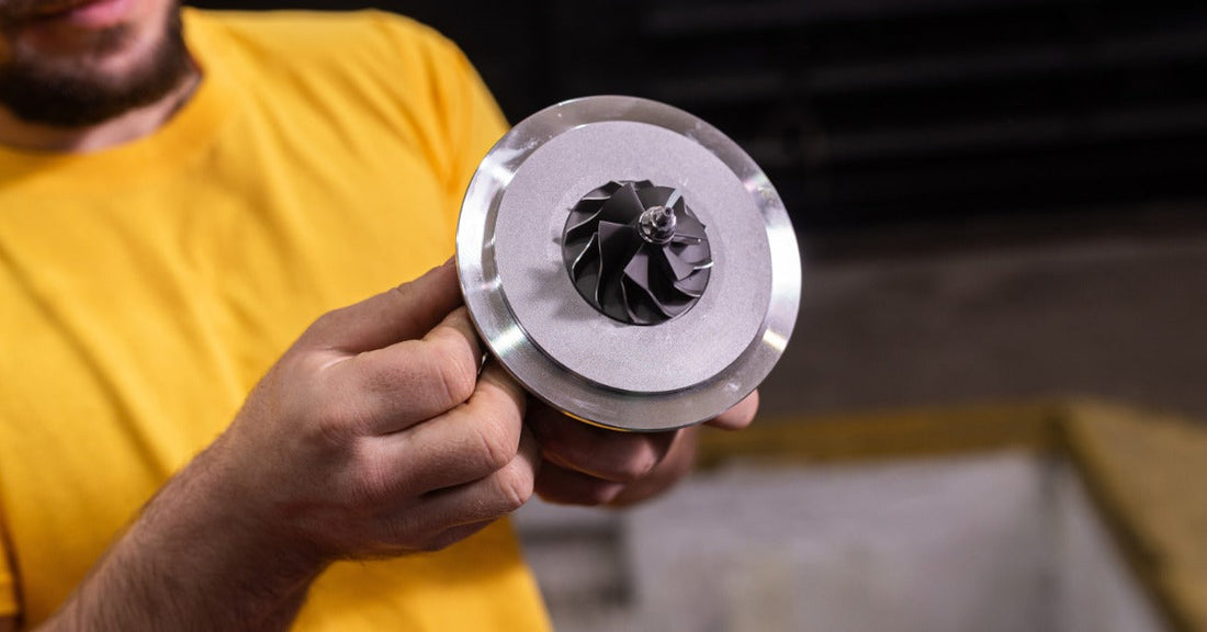 a person in a yellow shirt holding a round metal turbocharger cartridge with dark blades in a dim workshop.